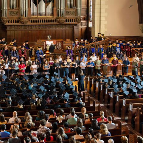 A large group of people sitting in a chapel. The people on stage are wearing different color graduation outfits while the people sitting at the pews are in traditional black graduation caps and gowns.
