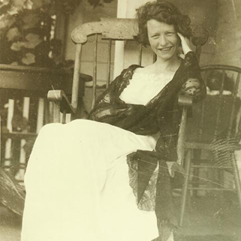 Edna St. Vincent Millay, smiling and wearing a long dress and lacy shawl, reclines in a rocking chair.