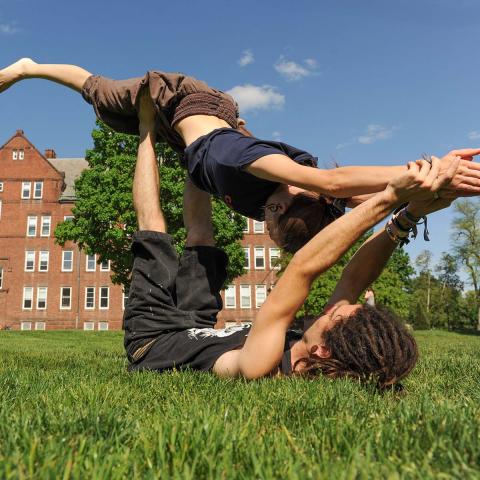 Two students practice yoga in the quad lying on the grass holding the other up in the air.