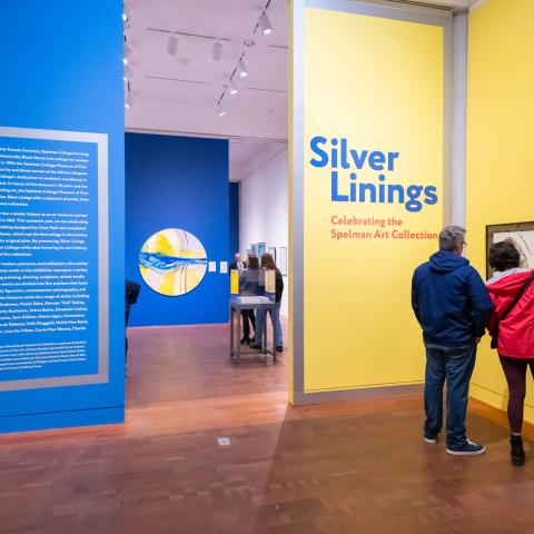 A room in the gallery with a blue wall on the left and yellow wall on the right. Two people are standing in front of a framed painting.