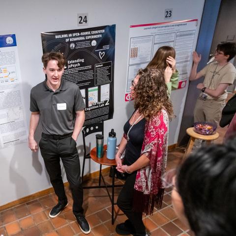 Groups of people standing in front of project posters on the wall while people listen.