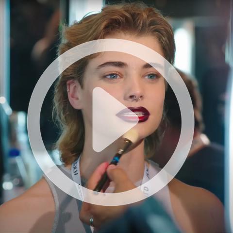 Mars Kubani getting makeup applied with video icon overlayed. 