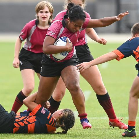 Person holding a rugby ball while others fight and struggle to stop, grab, hold her while playing rugby on a field.