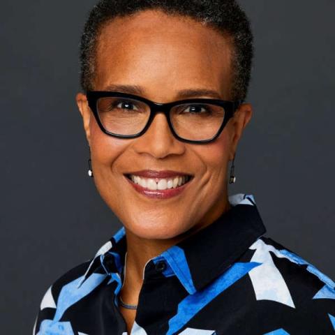 A person with short black curly hair, black-rimmed glasses, and a brightly patterned black and blue shirt smiles at the viewer.