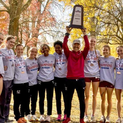 Group of smiling athletes on both sides of a coach in the center holding a trophy overhead.