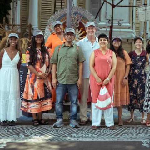 A group of people stand under a large straw roof, on a street with an intricate pattern painted on it. Behind them is a large stone building.