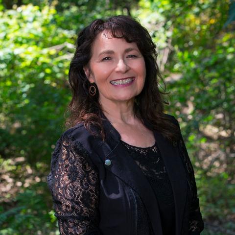 Pictured: Kimberly Blaeser. A person with a black shirt and brown hair smiling for the camera in front of green shrubbery. 