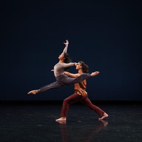 Two people on a dark stage with one person holding up the other in a split. 