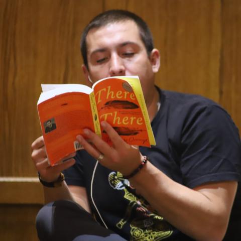 Seated person reading from a book titled, There There