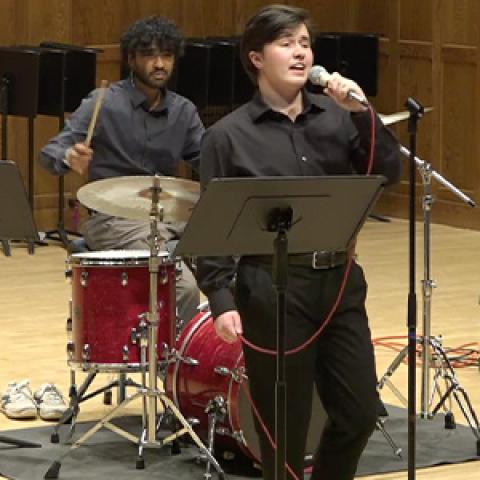 A closeup of two young men performing onstage, one singing into a mic and the other playing a drum set.