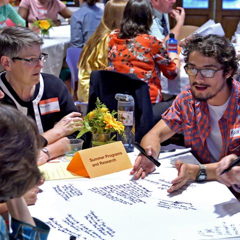 Group of people sitting at a table talking with notes on the table. The background room is filled with other round tables with people seated talking.