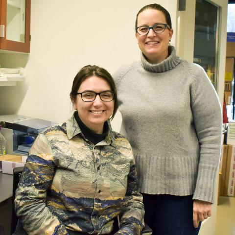 Morgan Stephens ’23 (left), sitting and wearing a printed collared shirt, and Visiting Scholar Elizabeth Thiele, standing and wearing a gray turtle neck sweater, in a research lab.