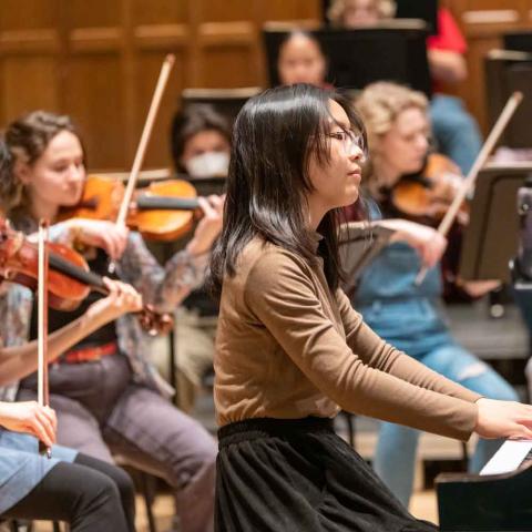 Sideview of people in an orchestra. One playing piano and the others playing violins..