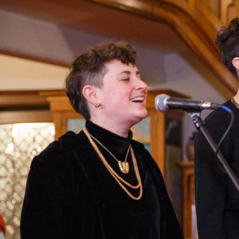 A person with short hair wearing a black long-sleeve shirt and gold necklace singing with a standing microphone.