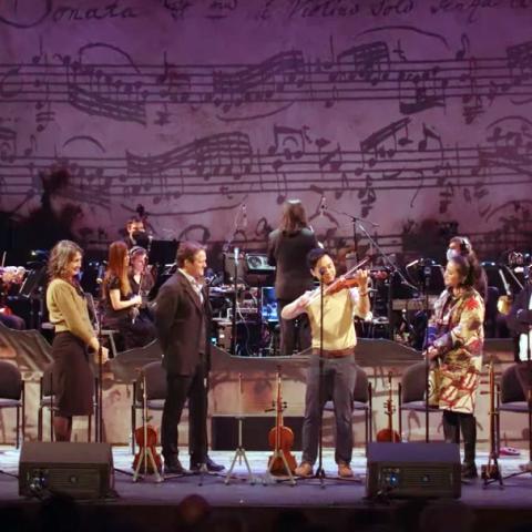 Multiple performers singing on stage in front of a symphony orchestra.