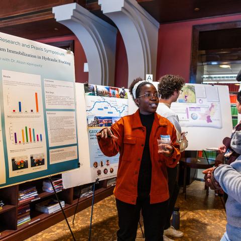 Person in a red sweater is pointing to a poster presentation on an easel while another person looks on.