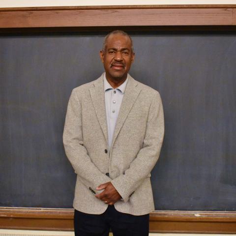Dr. Michael Gomez, wearing a light colored, collared shirt, beige jacket and black pants, standing in front of a blackboard.