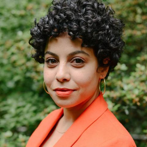 A person with short, black, curly hair and a bright orange suit jacket smiles at the viewer.