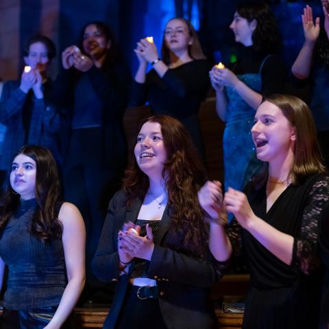 Performance of choir members standing, foreground row singing, background is a row of people holding a single candle each.