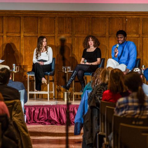 A panel of five people sitting in chairs on a stage with microphones speaking in front of an audience. 