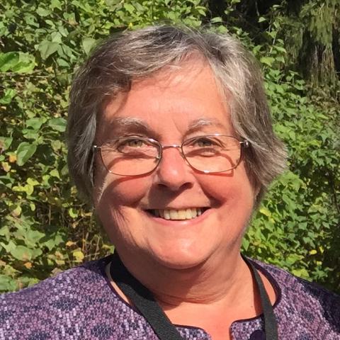 Person with glasses, purple shirt, and grey hair smiling in front of green shrubs outside.