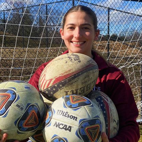 A person holding three soccer balls while standing outside on a soccer field in front of the goal/net.