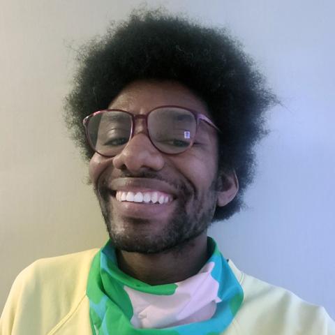 Headshot of a person with a vibrant smile wearing glasses, a yellow sweater and a scarf in teal, green, and white around the neck.