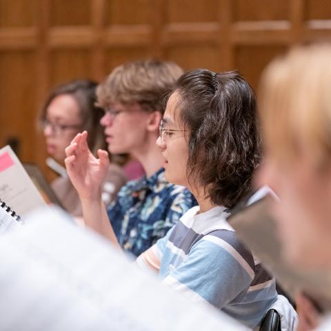 A row of singers in rehearsal showing four people, focused in the center on a person with glasses singing with her arm raised.