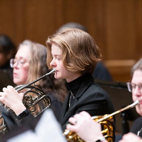 A performance, image focused on the center subject, a person with short straight hair playing the French horm.