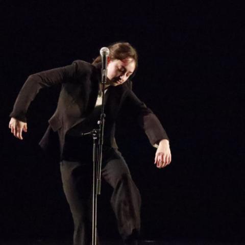 A person with long brown hair performs on a darkened stage in front of a microphone. The person is wearing all black clothing.