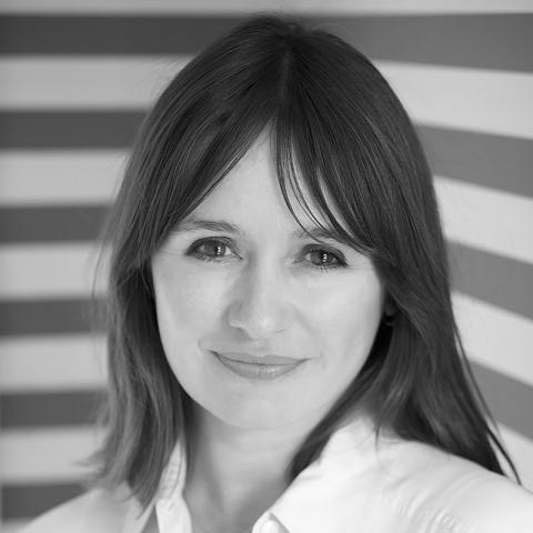 Black and white portrait of a person with shoulder-length hair, smiling slightly, wearing a white shirt, standing in front of a striped background. Photo credit: Robert Taylor.