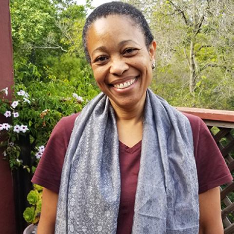 A portrait of Pam Harris standing outside on a deck with plants and trees all around.