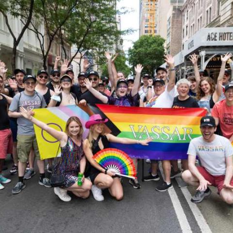 A group of smiling, waving people, some standing and some kneeling on the street in New York City, holding a rainbow banner with word "Vassar" on it.