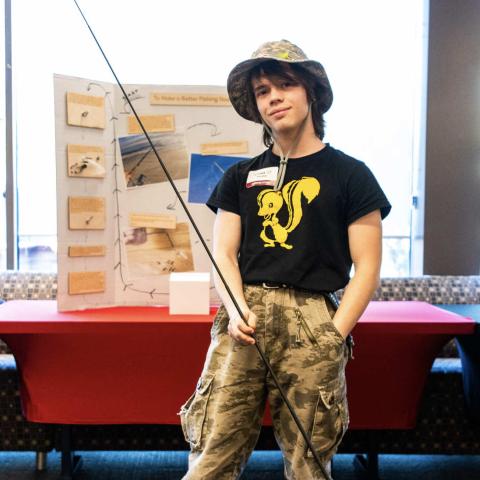 A person with long brown hair stands in front of a wall of large windows. The person is wearing a brimmed hat, camouflage pants, and a black T-shirt with a yellow cartoon drawing of a skunk on it. The person is holding a long fishing pole.