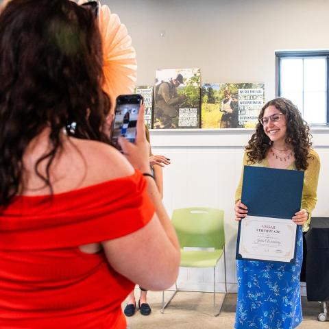 A person holding a diploma award smiling while another person holds a phone taking their picture.