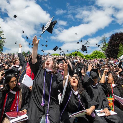 A large group of graduates in caps and gowns celebrate by throwing their mortarboards into the air at an outdoor graduation ceremony. The sky is bright with scattered clouds, and a crowd of onlookers fills the background.