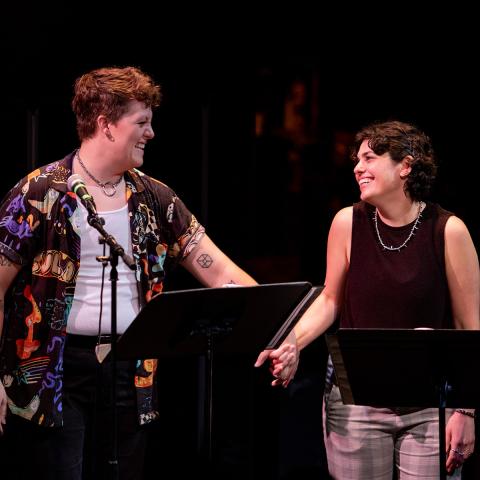 Two people on a dark stage smiling and looking at each other with microphones and podiums in front of them