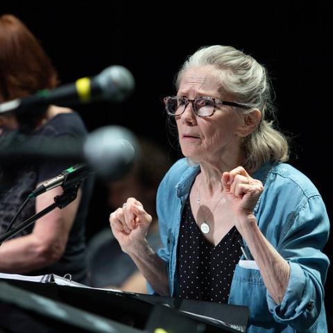 A person with long grey hair and glasses conducting on a dark stage in front of a podium and microphone.