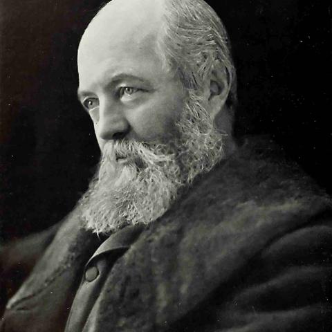 A historical portrait of Frederick Law Olmsted in 1893. Olmsted is a bald person with a long beard and a dark coat.