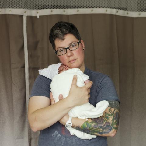 Photo taken in a hospital of nonbinary adult looking at the viewer while holding a newborn dressed in a white gown.