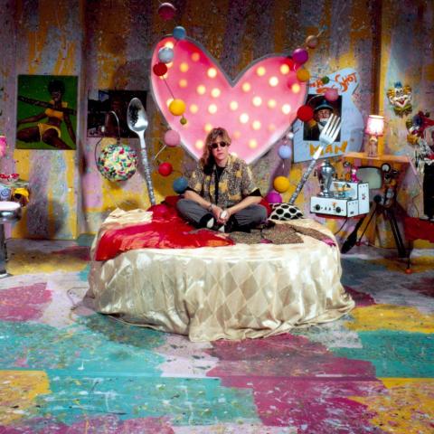 Allee Willis sits on a circular bed in the middle of a music video set, the walls are splatter painted and there is a heart with lights surrounding it on the wall behind her.