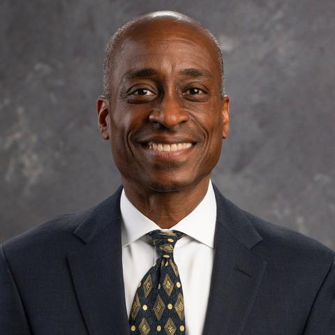 Portrait of a person smiling in a dark blue navy suit jacket, white shirt and blue and gold tie.