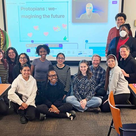 A group of people in a classroom posing in front of a screen with a presentation slide reading 'Protopians: we-imagining the future'.