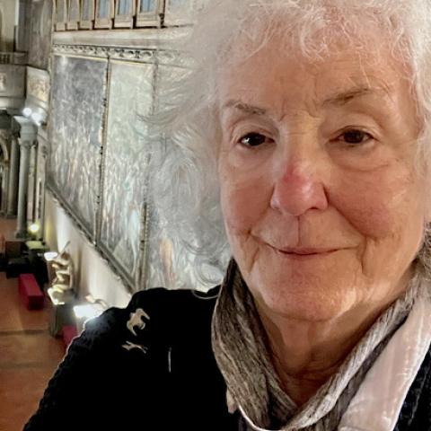 A person with fluffy grey hair stands in a large, ornate room with decorative moldings on the ceiling and walls.