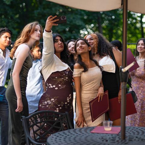 A group of young adults, dressed in elegant attire, gather under an outdoor umbrella for a selfie. 