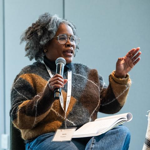 A person sitting on a stage speaking into a microphone to an audience.