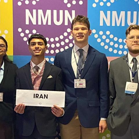 A group of smiling people standing in front of a backdrop with the acronym NMUN repeated: . A person in the middle is holding a sign that reads: Iran.
