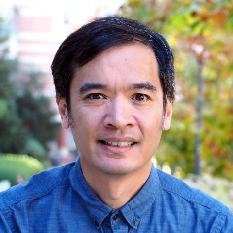 A smiling person wearing a blue shirt with short dark hair and blurred trees and partial building in the background.
