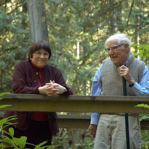 An elderly man and woman standing together on a wooden walkway in a lush, forested area, smiling and talking.