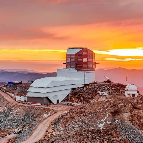 Vera C. Rubin Observatory sits atop a rocky mountain ridge at sunset, with vibrant orange and yellow hues lighting the sky.